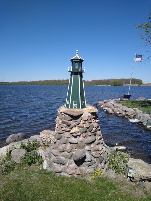 A green and white lighthouse on a stone base by a calm lake, with an American flag in the background and clear blue skies.