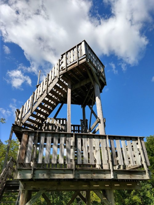 A wooden observation tower with stairs, set against a blue sky with fluffy clouds.