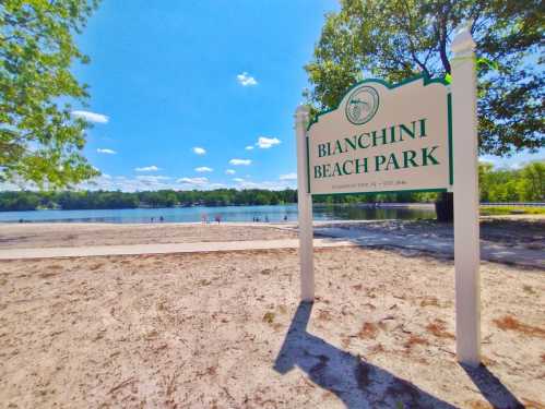 Sign for Bianchini Beach Park with a sandy beach and lake in the background, surrounded by trees and blue sky.