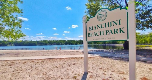 Sign for Bianchini Beach Park by a sandy beach and calm water, with trees and blue sky in the background.