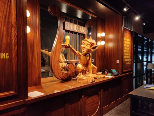 A wooden sculpture of a playful goblin holding a drink, set against a stylish bar backdrop with warm lighting.