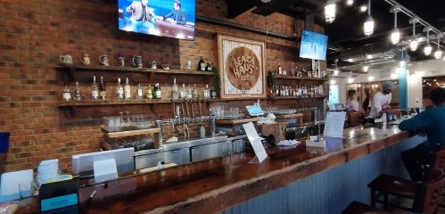 A bar with a wooden counter, shelves of liquor, and two TVs displaying sports in a cozy, rustic setting.