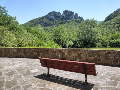 A red bench on a stone patio overlooks a lush green landscape and rocky mountains under a clear blue sky.