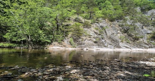A serene riverbank with smooth stones, surrounded by lush green trees and rocky cliffs reflecting in the calm water.