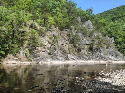 A serene river scene with rocky cliffs and lush greenery, reflecting in calm water under a clear blue sky.