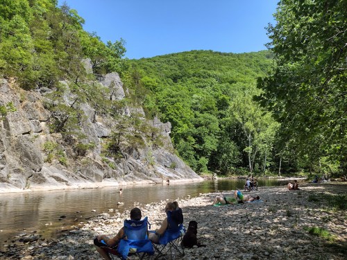 People relax by a riverbank surrounded by lush greenery and rocky cliffs on a sunny day.