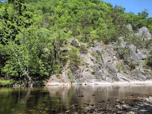 Two people wade in a calm river beside a rocky shore, surrounded by lush green trees and a clear blue sky.