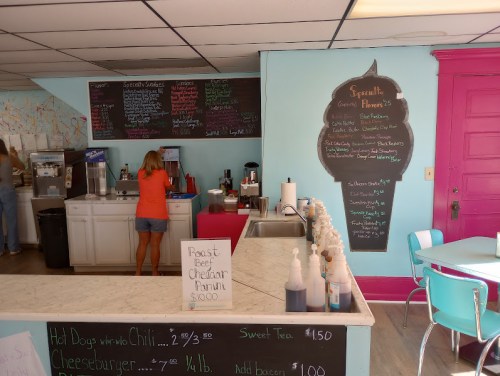 A colorful café interior with a menu board, counter, and seating area. A person is preparing food behind the counter.