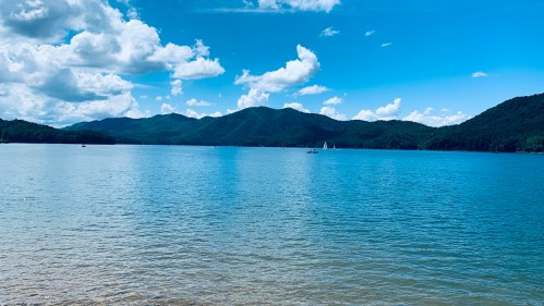 A serene lake surrounded by mountains under a bright blue sky with fluffy clouds.