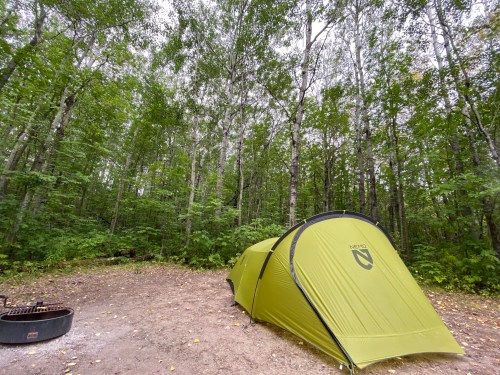 A green tent set up in a forest clearing, surrounded by tall trees and a fire pit nearby.