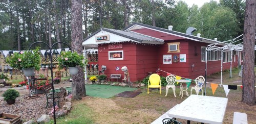 A cozy red building with an "Open" sign, surrounded by flowers and outdoor seating in a wooded area.