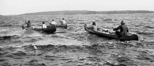Two canoes with people paddling across a wavy body of water, surrounded by trees in the background.