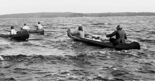 Black and white photo of several people in canoes paddling across a lake with trees in the background.