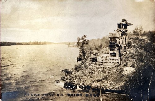 Historic photo of a watermill by a river, surrounded by trees and rocky shoreline, with a cloudy sky above.
