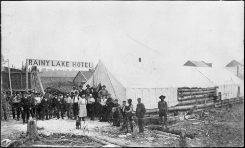 A historical black-and-white photo of a group of people in front of the Rainy Lake Hotel, with tents and log structures.