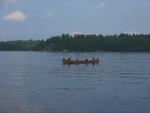 A canoe with four people paddling on a calm lake, surrounded by lush green trees and a cloudy sky.