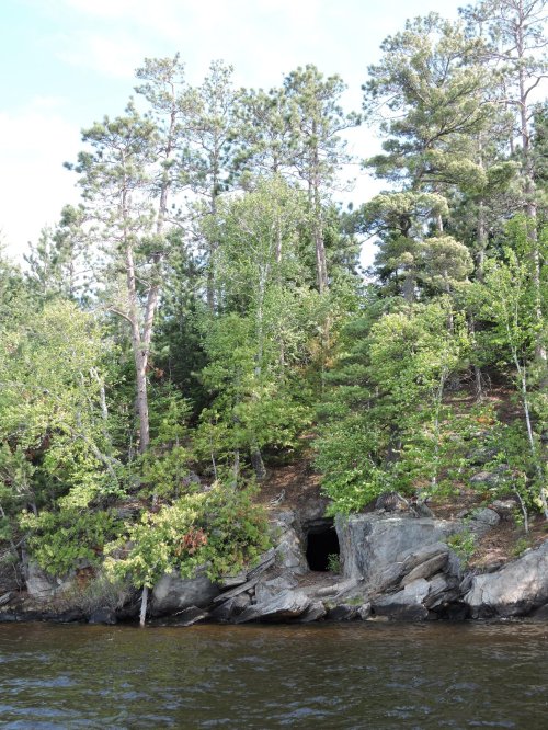 A rocky shoreline with dense green trees and a small cave entrance visible among the foliage.