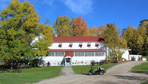 A white building with a red roof surrounded by colorful autumn trees, with a golf cart parked nearby on a gravel path.