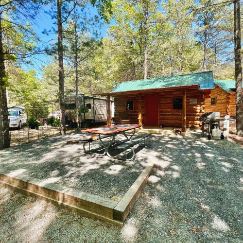 A cozy log cabin surrounded by trees, featuring a picnic table and grill in a gravel area.