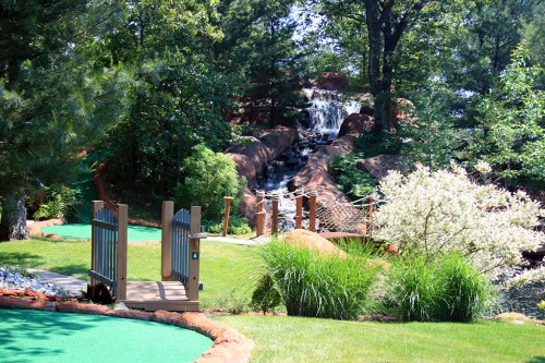 A scenic mini-golf course featuring a waterfall, lush greenery, and a wooden bridge surrounded by trees.