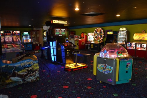 A colorful arcade filled with various games, including skee-ball and a cyclone machine, under dim lighting.