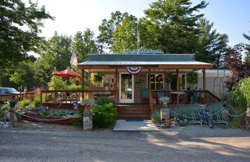 A charming, rustic building with a porch, surrounded by greenery and outdoor seating, set in a peaceful rural area.