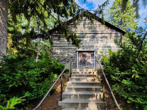 A rustic wooden building with a staircase leading up, surrounded by greenery and trees under a blue sky.