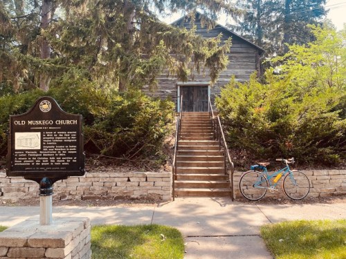 A historic wooden church with stairs leading up, beside a sign and a parked bicycle on a sunny day.