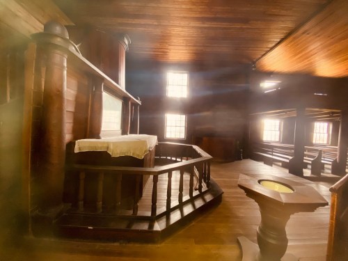 Interior of a wooden chapel with a simple altar, benches, and soft light filtering through the windows.