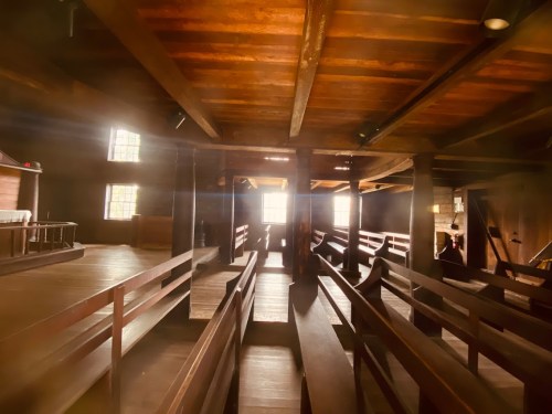 Interior of a rustic wooden building with benches, beams, and soft light streaming through windows.