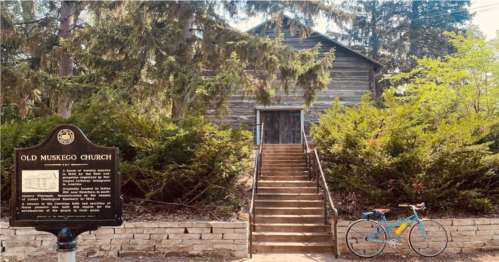 A historic wooden church with a sign in front, surrounded by greenery and a bicycle parked nearby.
