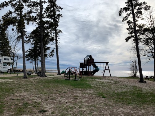 A playground with slides and swings near a lake, surrounded by trees and a cloudy sky.