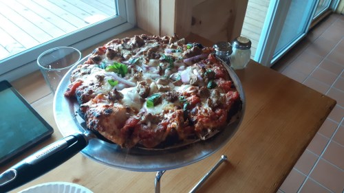 A freshly baked pizza topped with cheese, meat, and green herbs, served on a wooden table near a window.
