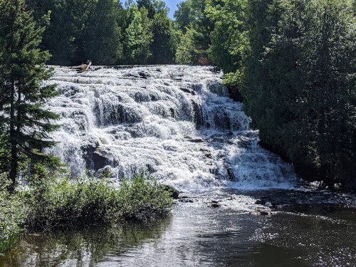 A cascading waterfall flows over rocks, surrounded by lush green trees and a calm river below.