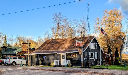 A rustic building with a Coca-Cola sign, an American flag, and a moose statue, surrounded by autumn trees.