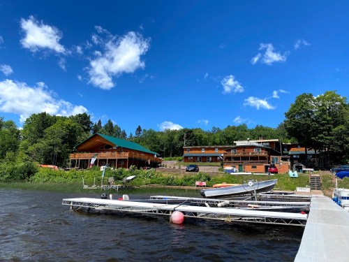 A scenic view of a river with boats docked, surrounded by green trees and a lodge under a blue sky with fluffy clouds.