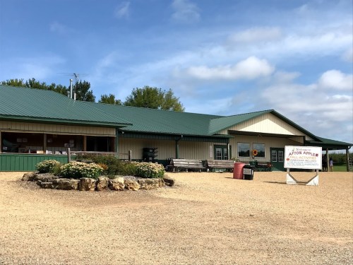 A rural building with a green roof, surrounded by gravel and flowers, featuring a sign for an apple orchard.
