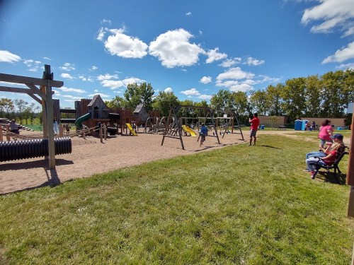 A sunny playground with swings, slides, and children playing, surrounded by green grass and trees under a blue sky.