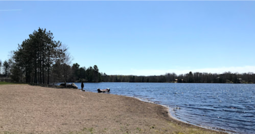 A serene lakeside scene with a sandy shore, trees, and a clear blue sky. A person stands near the water's edge.