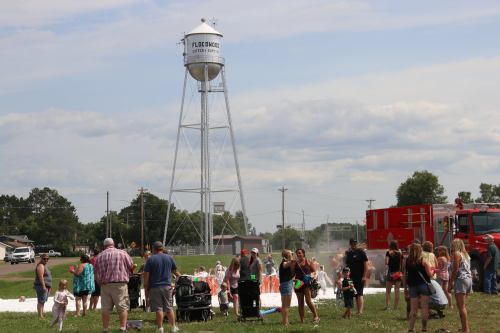A crowd enjoys a summer event with a water tower in the background, foam on the ground, and a fire truck nearby.