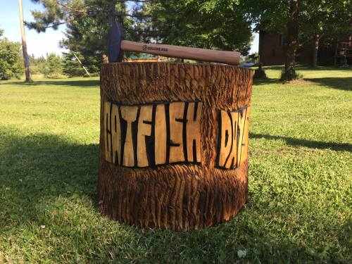 A wooden stump with "Catfish Day" carved into it, topped with a hatchet, set on a grassy area with trees in the background.