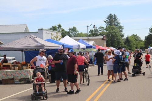 A lively street market scene with people browsing stalls, chatting, and enjoying the day under colorful tents.