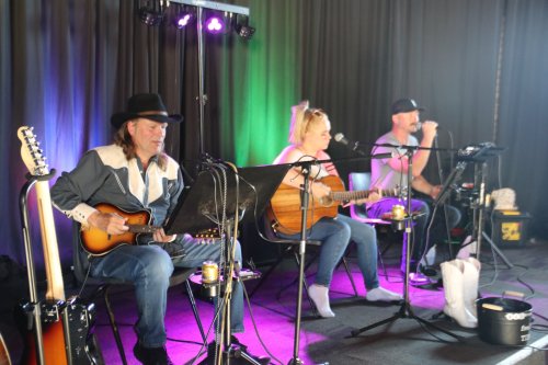 Three musicians perform on stage with guitars, singing into microphones, under colorful lights.