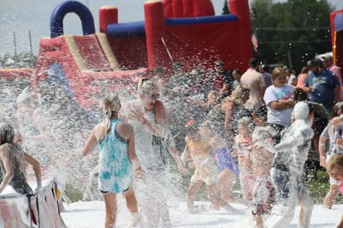 A lively outdoor scene with children and adults playing in foam, surrounded by a bouncy castle and a crowd enjoying the festivities.
