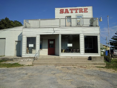 A weathered building with "SATTRE" sign, featuring large windows and a staircase, set against a clear blue sky.