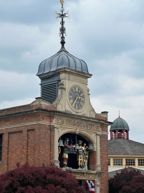 Clock tower with statues and a weather vane, featuring a historical inscription, against a cloudy sky.