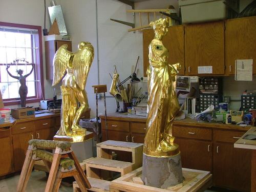 Two golden statues in a workshop, one with wings and the other draped in fabric, surrounded by tools and materials.