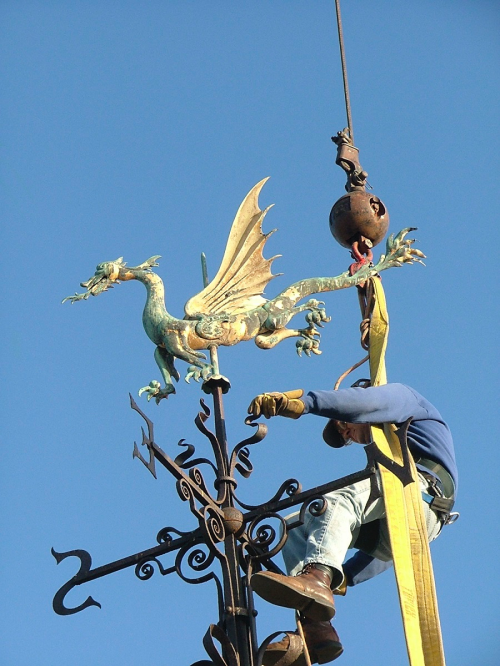 A worker adjusts a dragon weathervane atop a building against a clear blue sky.