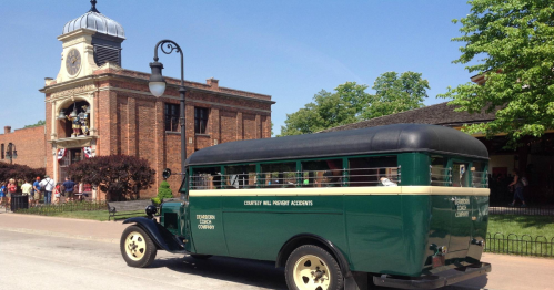 A vintage green and black bus parked in front of a historic brick building with a clock tower on a sunny day.