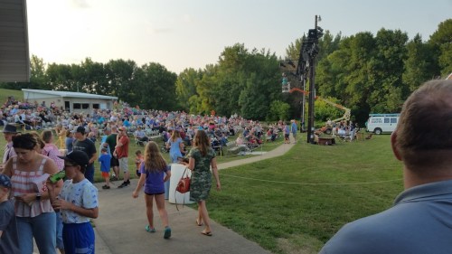 A large crowd gathers on a grassy area for an outdoor event, with people seated and standing, enjoying the atmosphere.